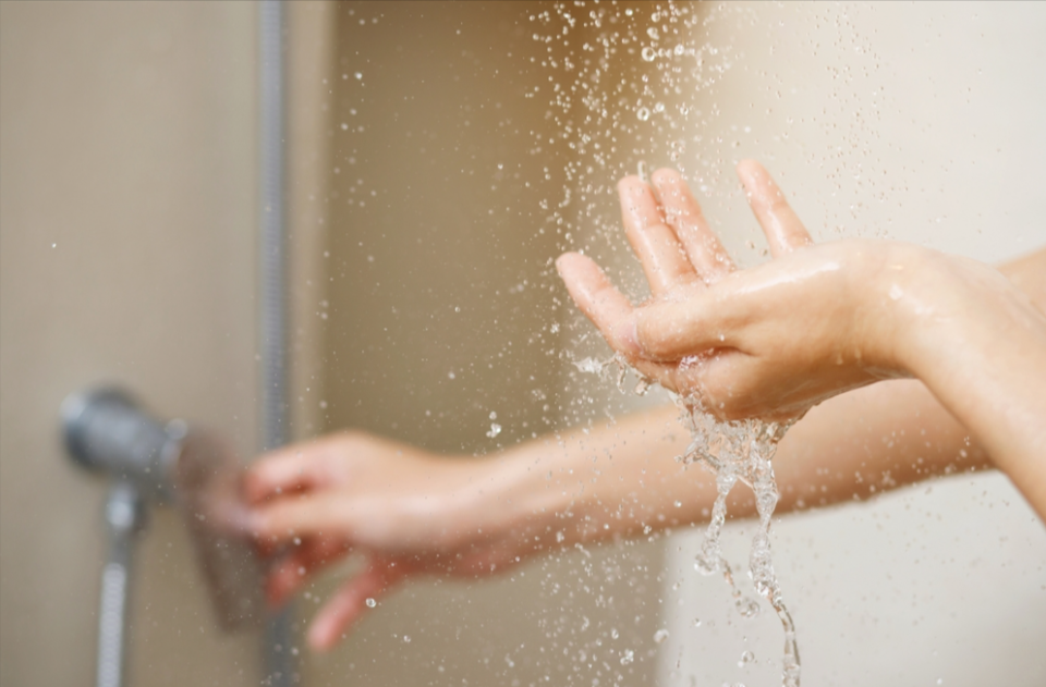 A person holds their hand under running shower water while adjusting the shower handle with their other hand.