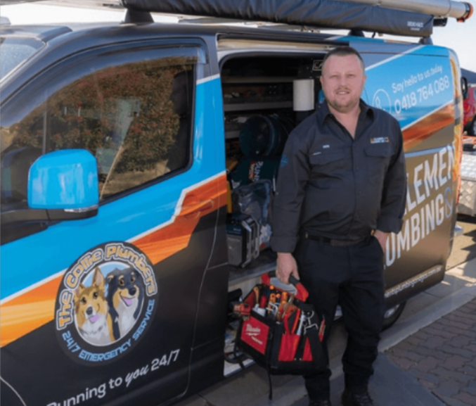 A plumber stands next to a van with “The Canine Plumbers” logo, holding a red tool bag. The van has a colorful wrap featuring two cartoon dogs and plumbing equipment is visible inside.